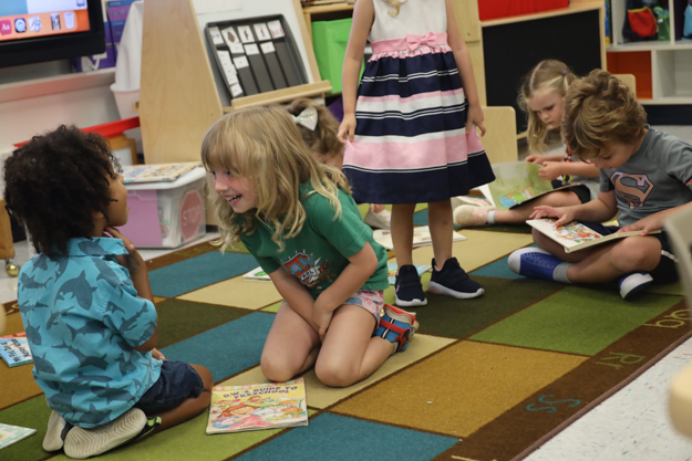 Two young children laugh together while other children in the background read a book with a teacher.